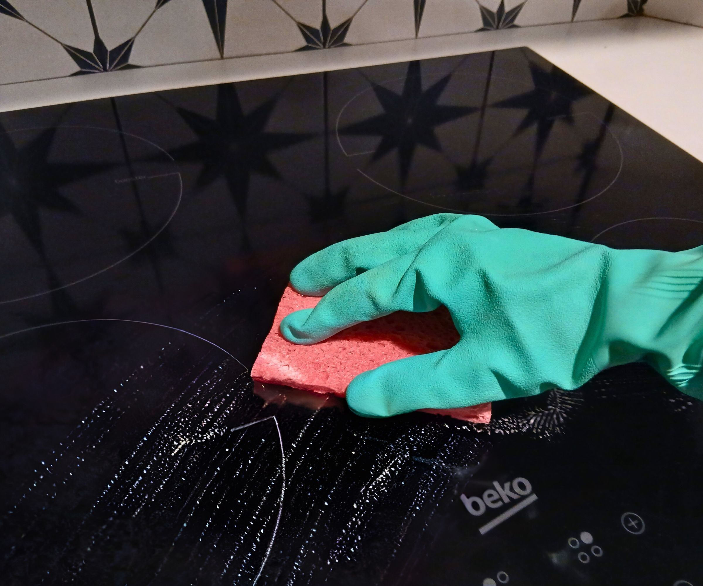 A gloved hand moves a pink sponge across a glossy black induction hob, lifting away streaks of cleaner.