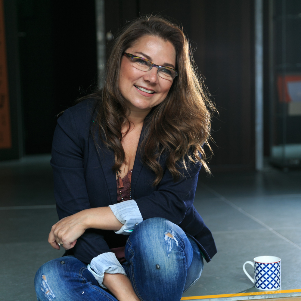 woman with long brown hair and glasses sat on tiled floor wearing navy jacket and jeans