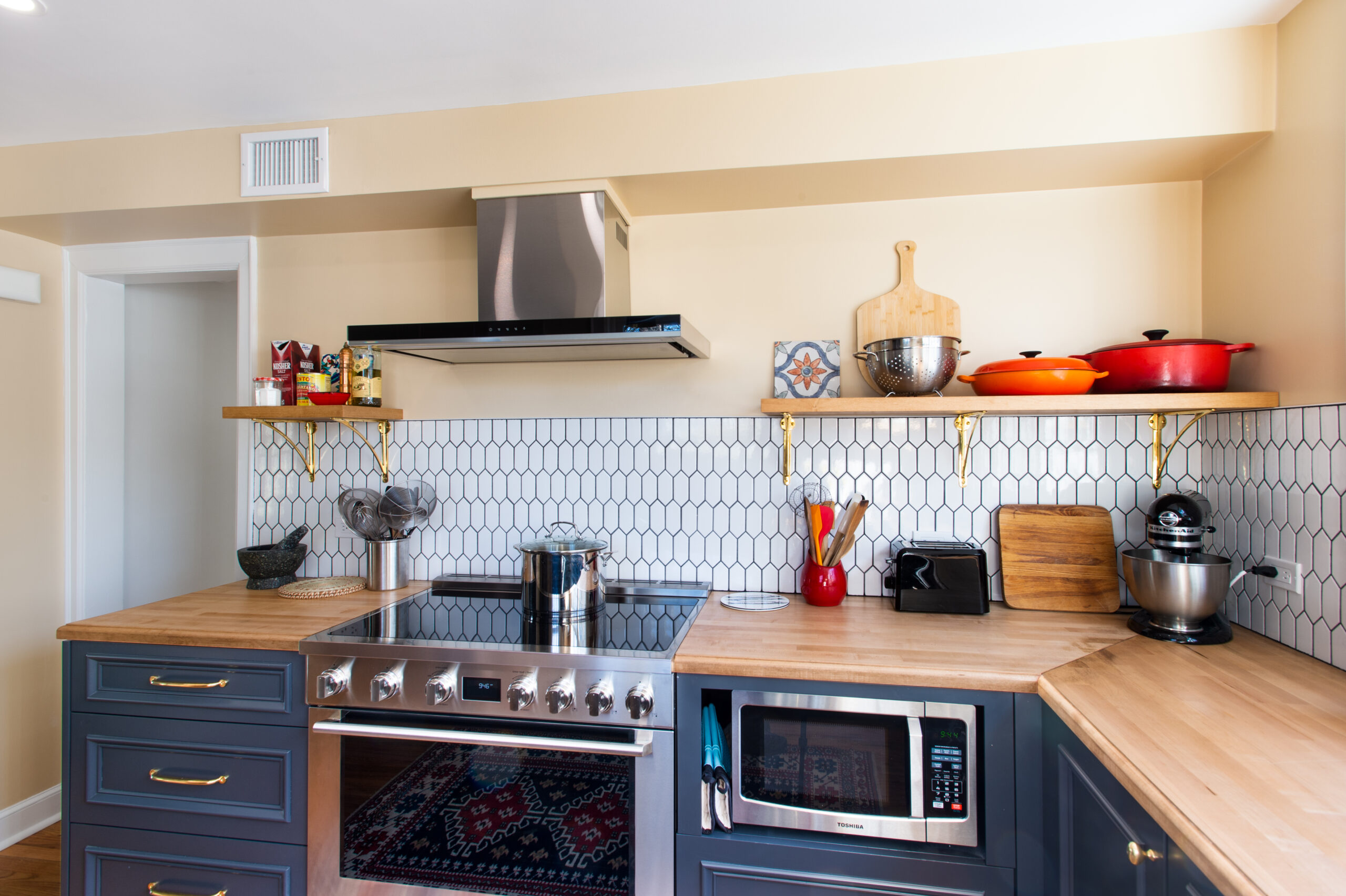 Corner kitchen with blue base cabinets, open wood shelves, white patterned tile and continuous butcher block countertops for prep and small appliances