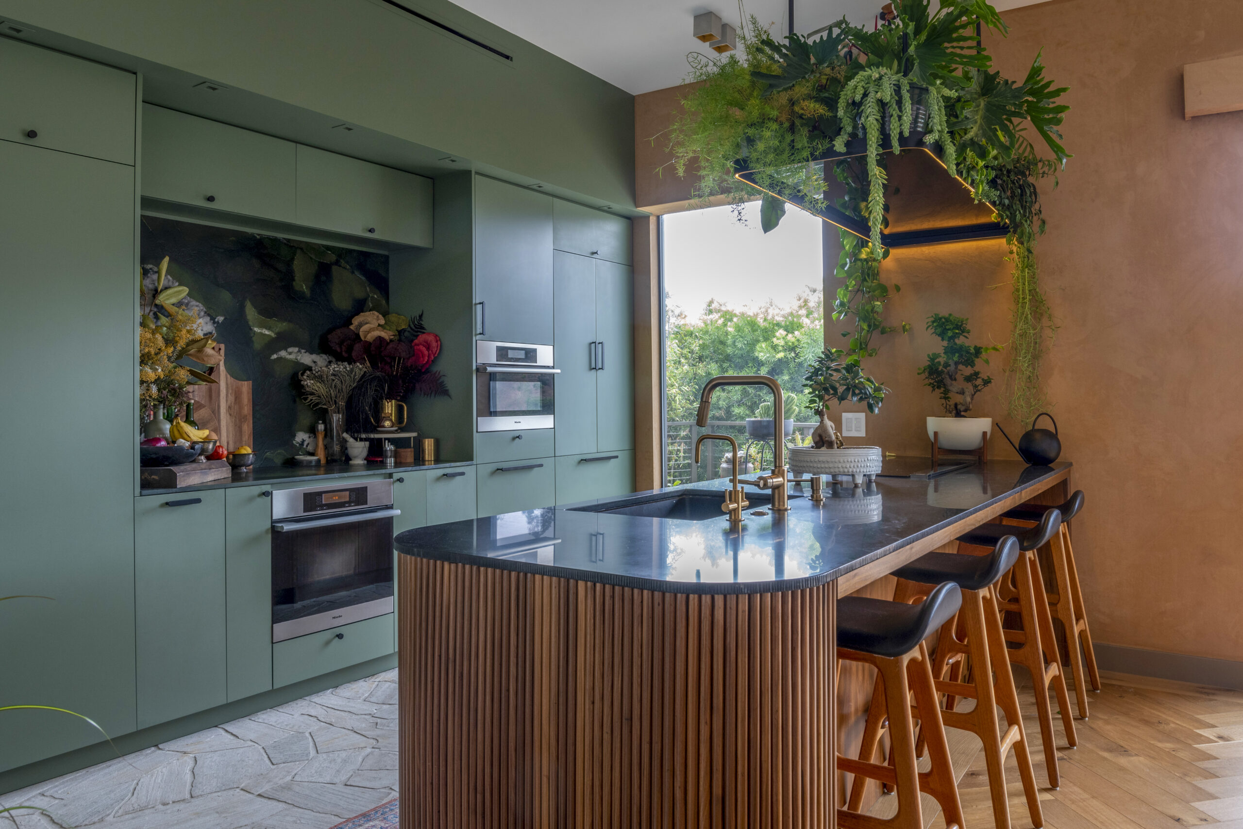 Sage green cabinetry and warm wood textures pair with lush greenery for a dramatic, nature-inspired modern kitchen.