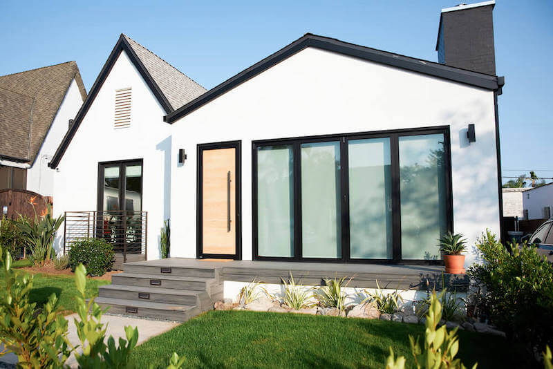 Scandi-modern front exterior with white stucco siding, black trim windows, light wood front door, wide sliders, new steps, and fresh landscaping.