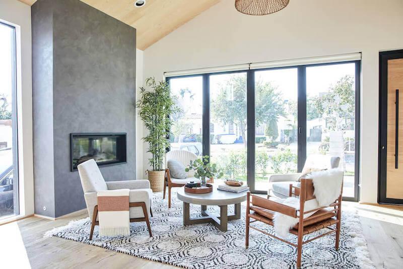 Sunlit sitting area with tall gray fireplace column, layered rugs, midcentury inspired chairs, and expansive glass doors facing the yard.