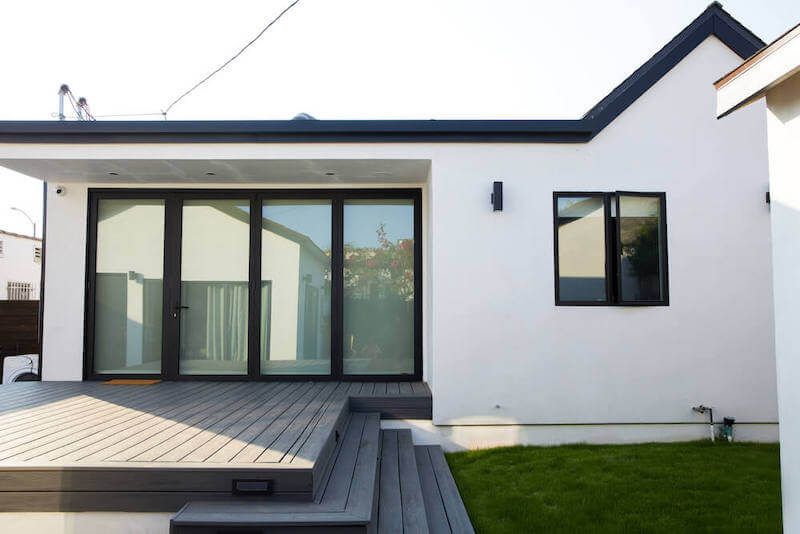 Backyard view of Scandi-modern home with white stucco walls, dark framed sliding glass doors, raised composite deck, and small lawn area.