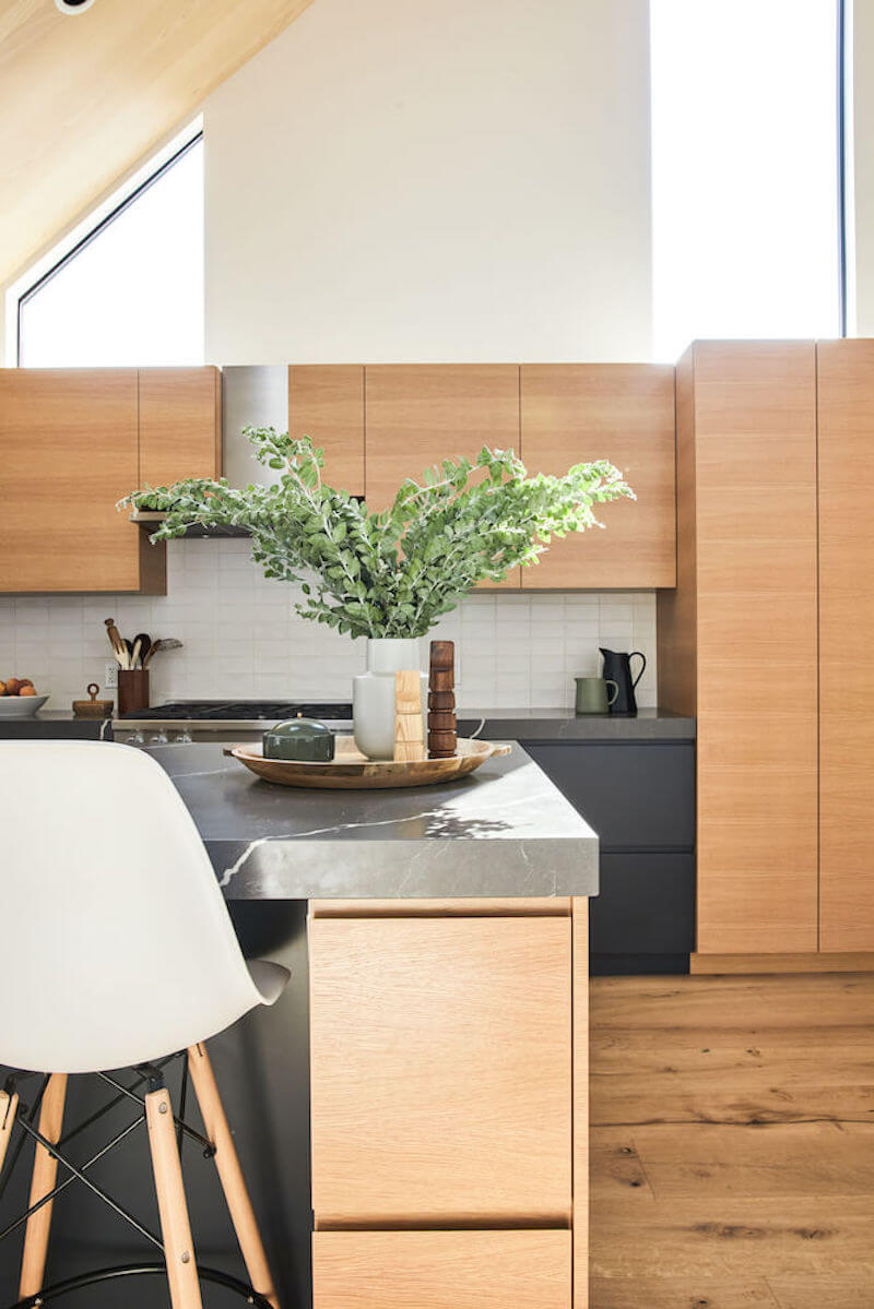 Detail of kitchen island with stone waterfall counter, wood drawer fronts, and styled tray, framed by full height cabinets and light walls.