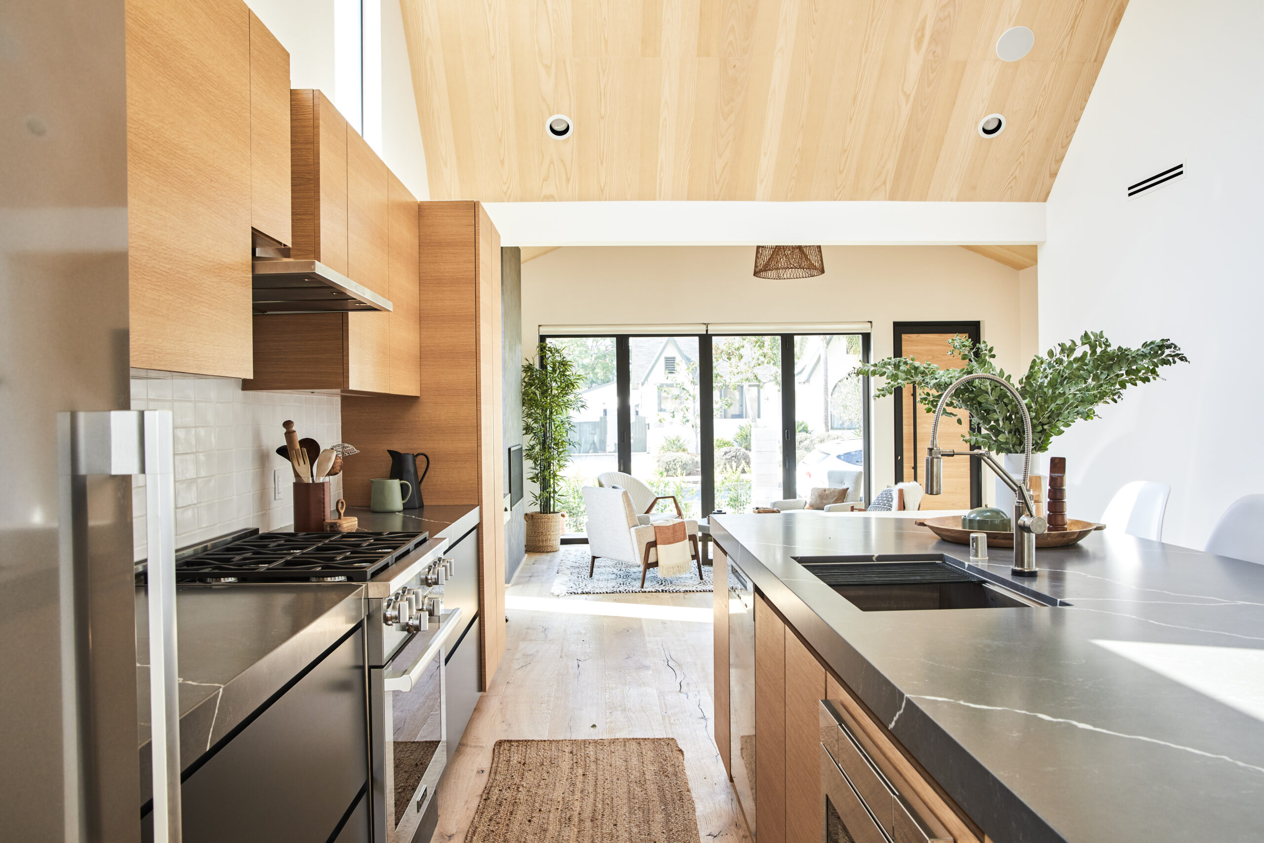 Kitchen view toward living space showing long stone island, wood cabinetry, stainless range, and sliding glass doors that open to the yard.