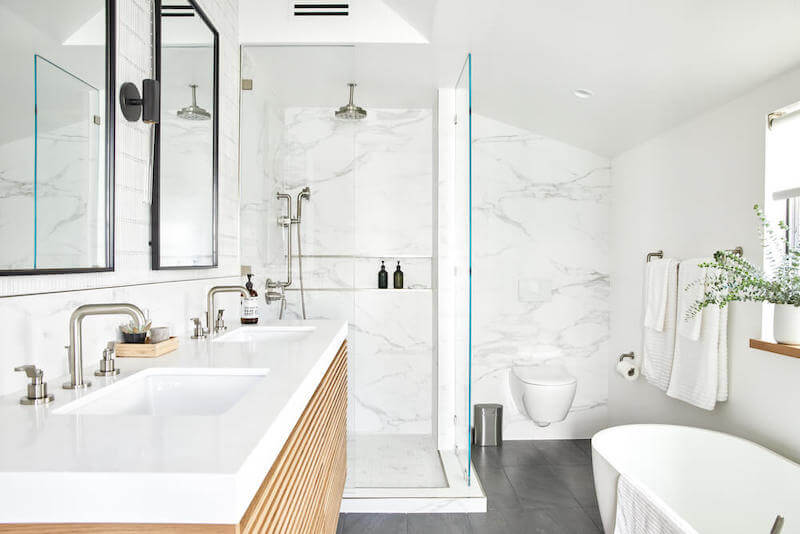 Bright main bathroom with double vanity, walk in marble tile shower, wall hung toilet, freestanding soaking tub, and large format gray floor tile.