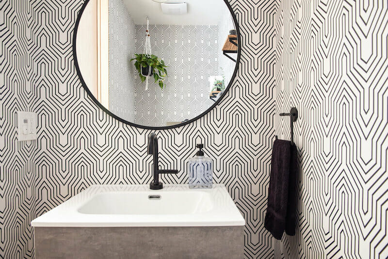 Small powder room with bold black and white geometric wallpaper, round mirror, compact vanity with integrated sink, and matte black faucet.