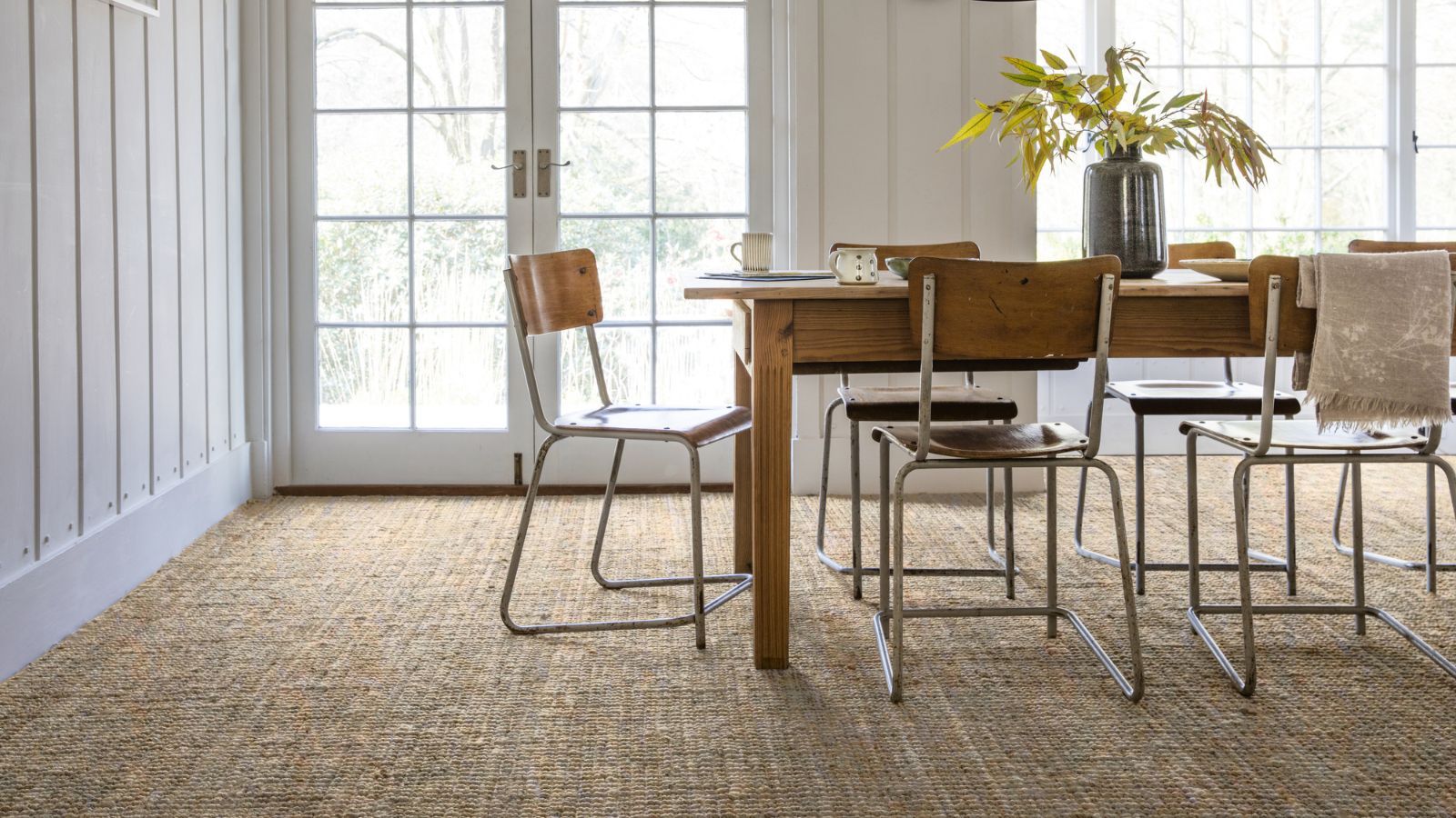 A light-filled dining room featuring a rustic wooden table and vintage metal chairs on a textured woven carpet. Large French doors flood the space with natural light, highlighting the room’s relaxed, natural style.