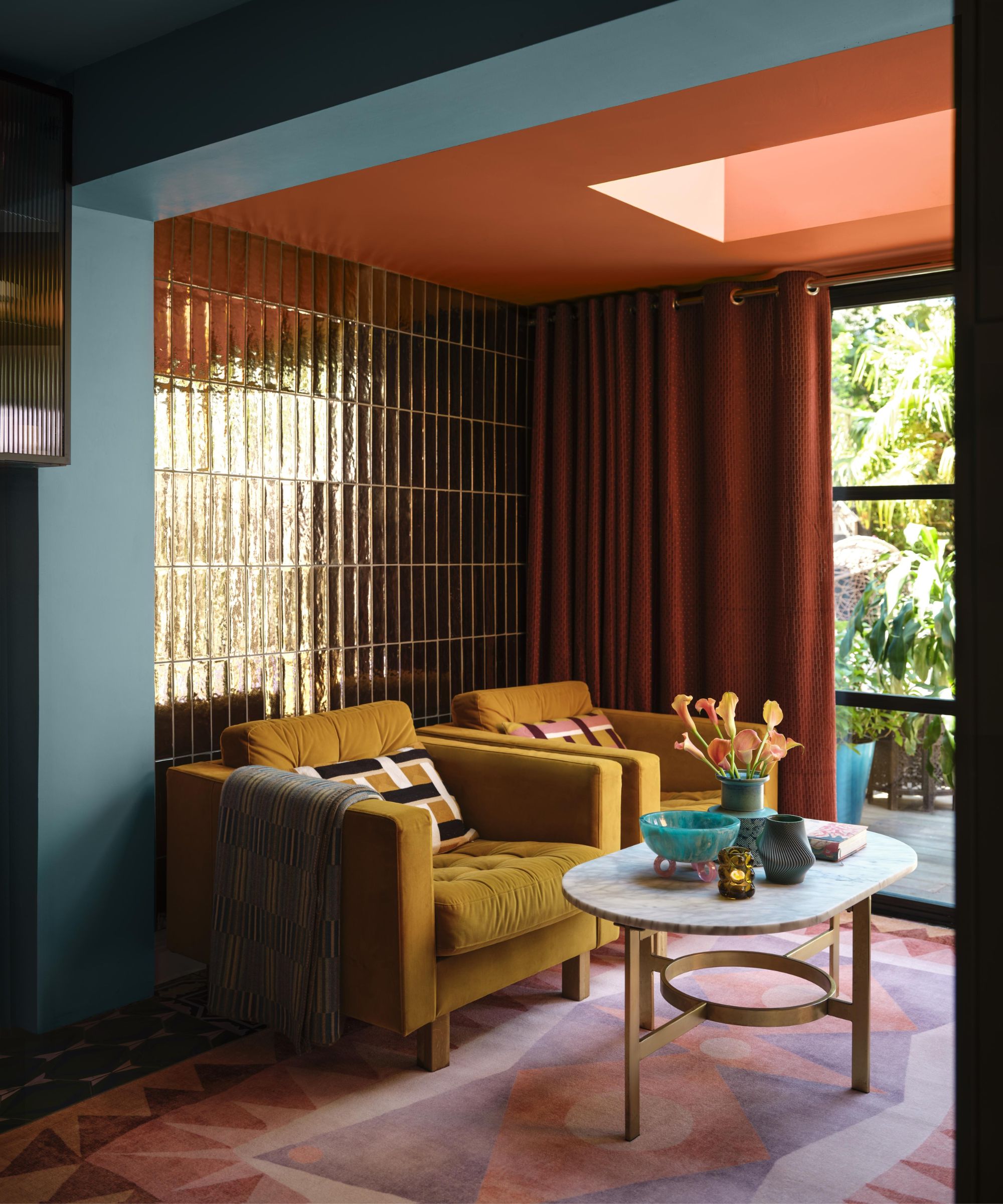 Colourful living room with mustard velvet armchairs, metallic gold wall tiles, terracotta curtains and a marble coffee table styled with vibrant accessories.