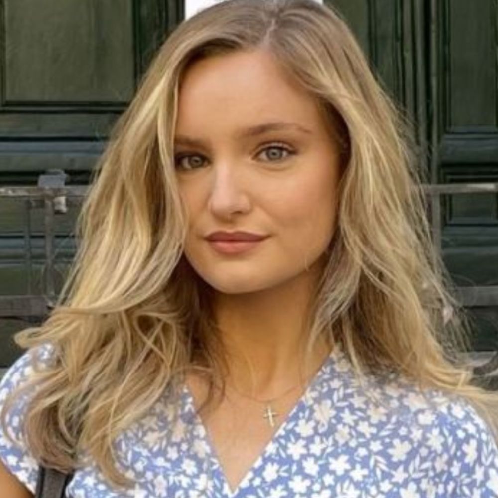 A portrait of a woman with long blonde hair wearing a blue floral dress and a delicate cross necklace, standing outdoors in front of dark green wooden doors