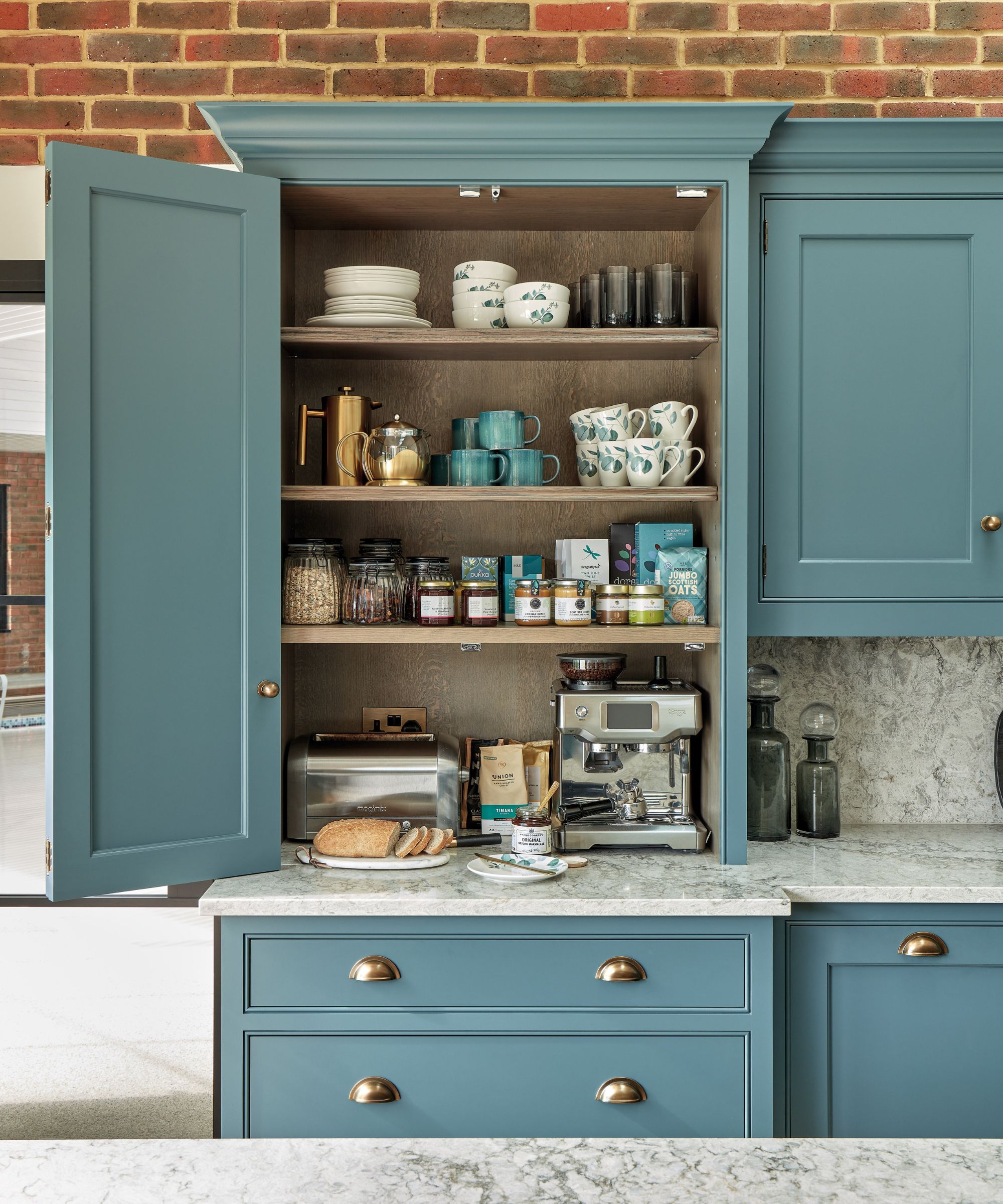 A built-in blue kitchen cupboard styled as a coffee station, with open shelves displaying mugs, jars, coffee accessories and an espresso machine. Brass handles, a marble worktop and exposed brick wall add contrast, while the open cabinet doors showcase practical yet decorative storage.