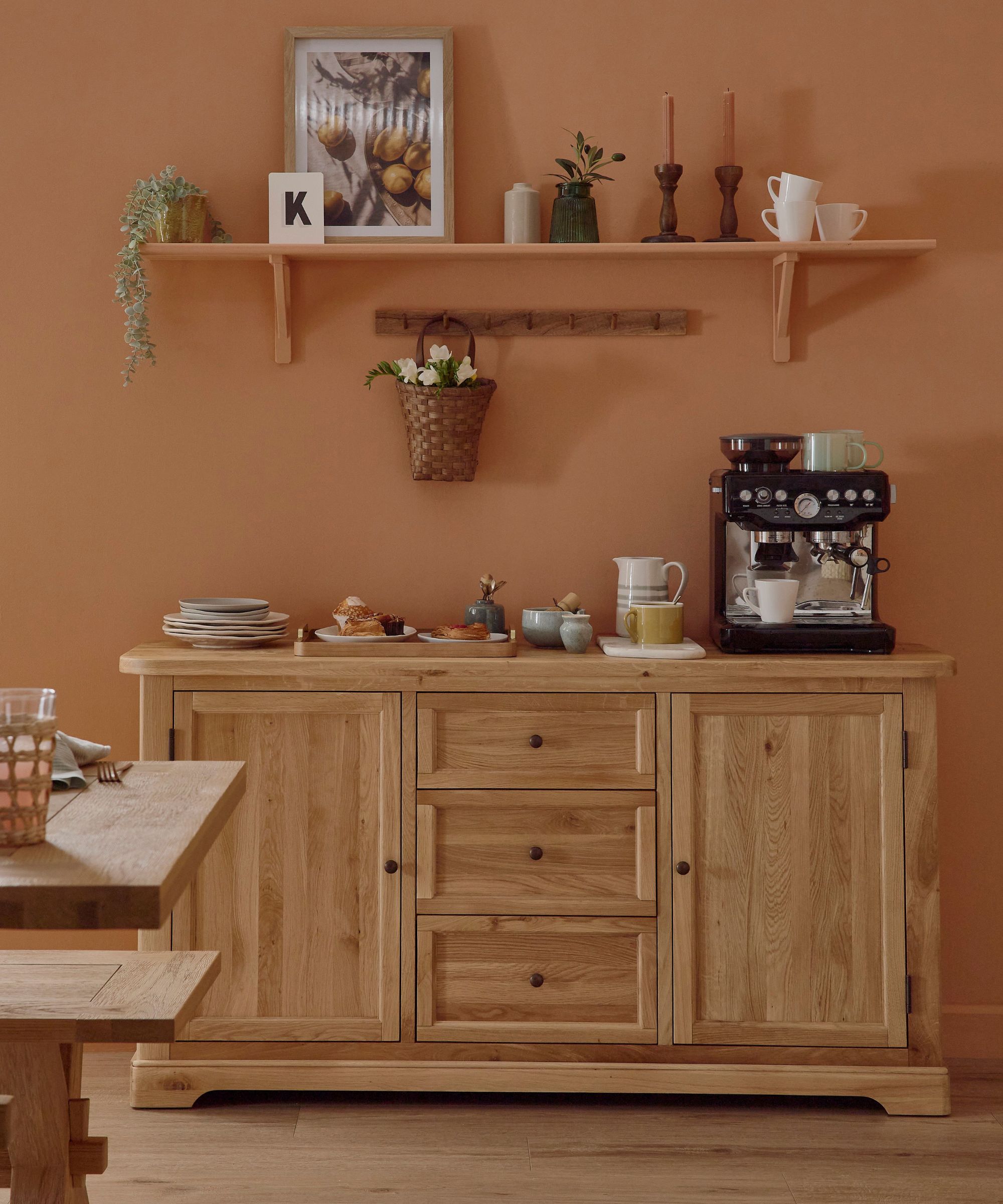 A warm, rustic coffee station styled against a terracotta wall. An oak sideboard holds a coffee machine, mugs and pastries, while simple shelves above display art, candles and greenery, giving the dining space a relaxed, homely feel.