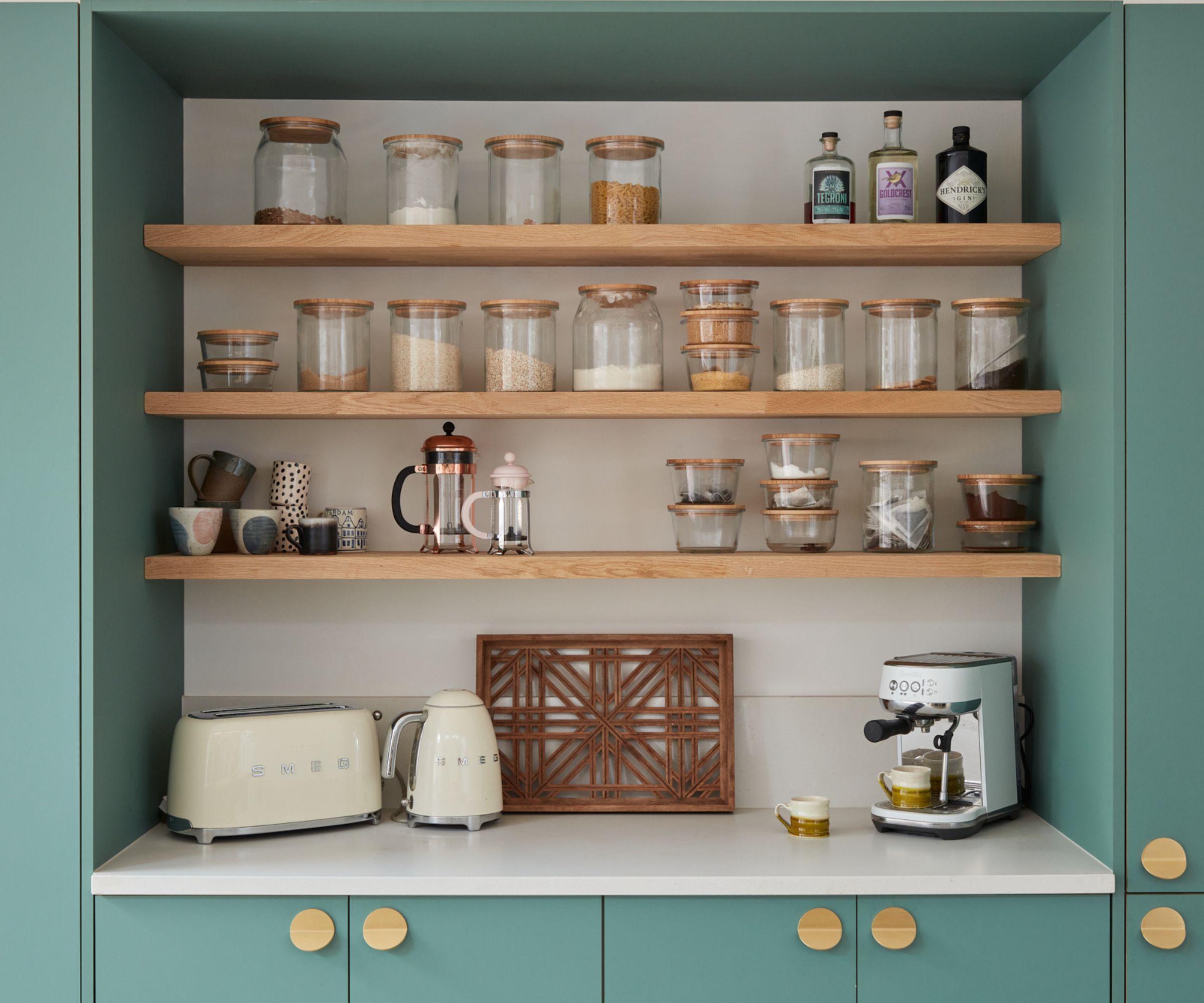 A beautifully organised coffee and breakfast station with sage-green cabinetry, open oak shelves and glass jars filled with dry goods. A pastel kettle, toaster and compact espresso machine sit on a clean white worktop, creating a calm, practical kitchen nook.
