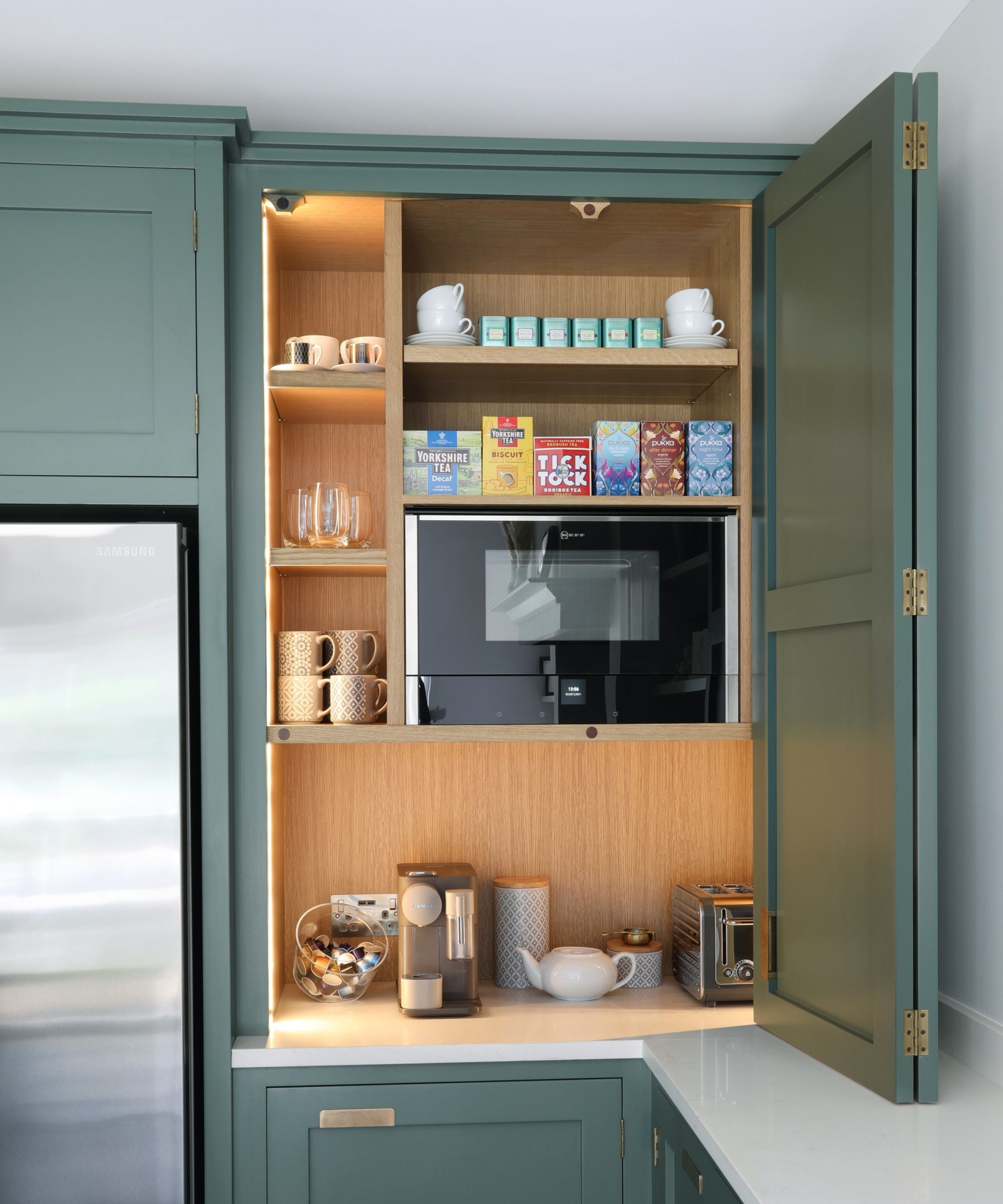 An elegant kitchen cupboard opens to reveal a well-organised breakfast and drinks station with illuminated wooden shelving. Tea boxes, cups, a microwave, mugs, a coffee machine and toaster are neatly arranged within muted green cabinetry and a clean, modern worktop.