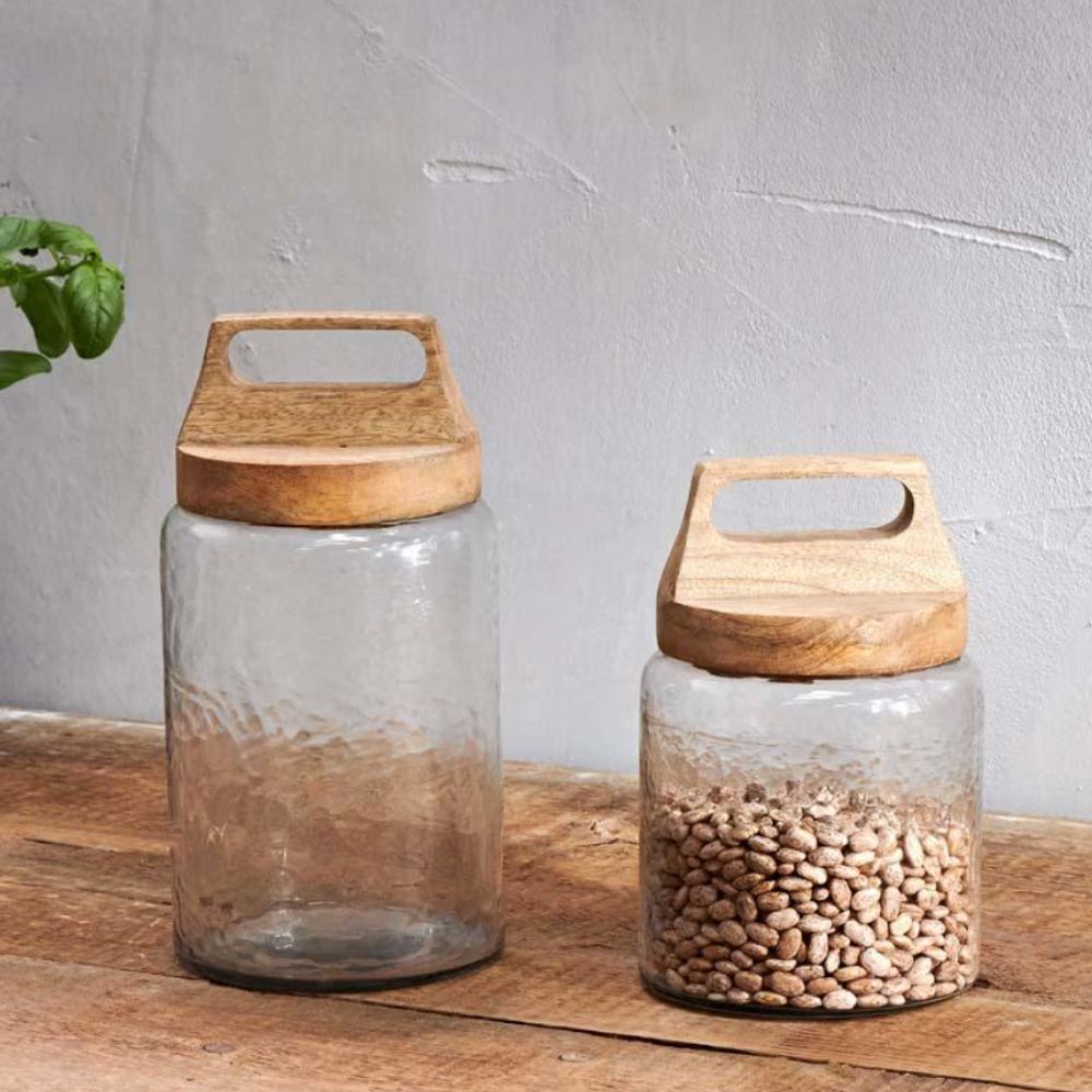 Set of two clear glass kitchen storage jars with natural wooden lids, one filled with dried beans and one empty, displayed on a rustic wooden table.