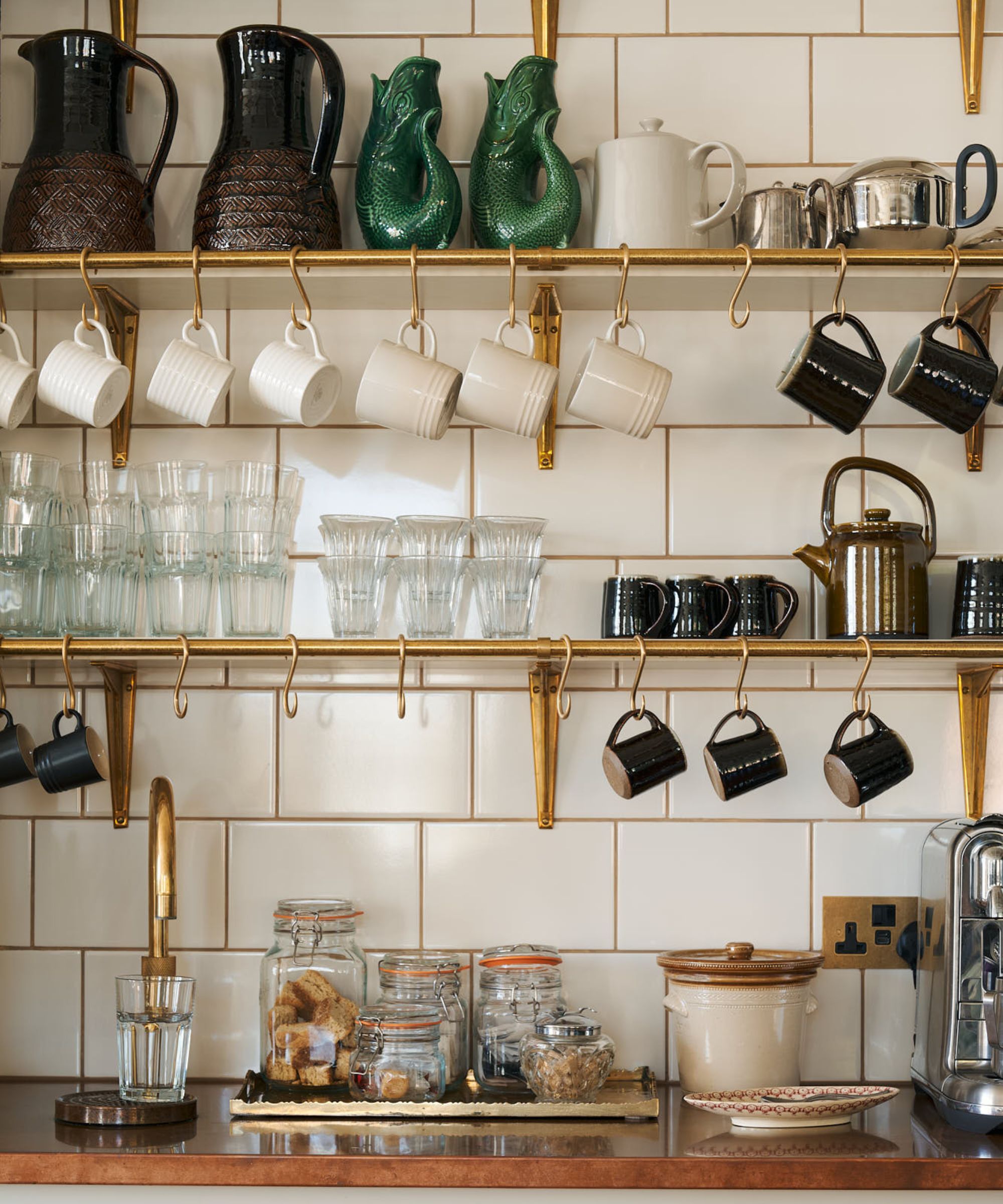 A neatly organised home coffee station with brass wall-mounted shelves and hooks holding ceramic mugs and glass tumblers. Below, a countertop displays clear storage jars filled with coffee and biscuits, a traditional kettle, and a stainless steel espresso machine.