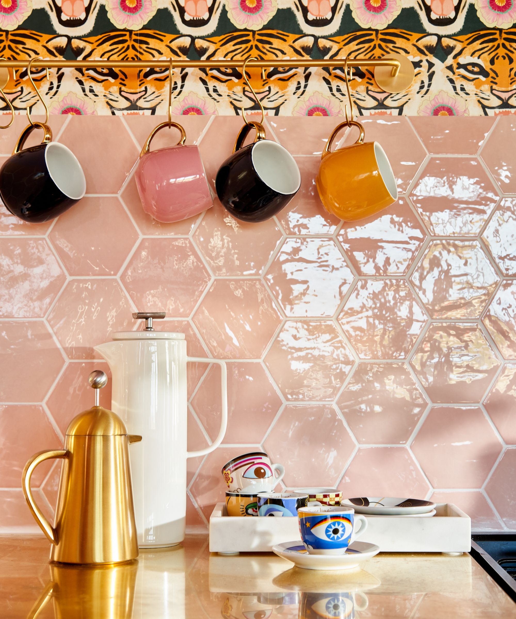 Colourful coffee station with hanging mugs on brass rail, pink hexagon tiles, patterned wallpaper and gold coffee pot on countertop.
