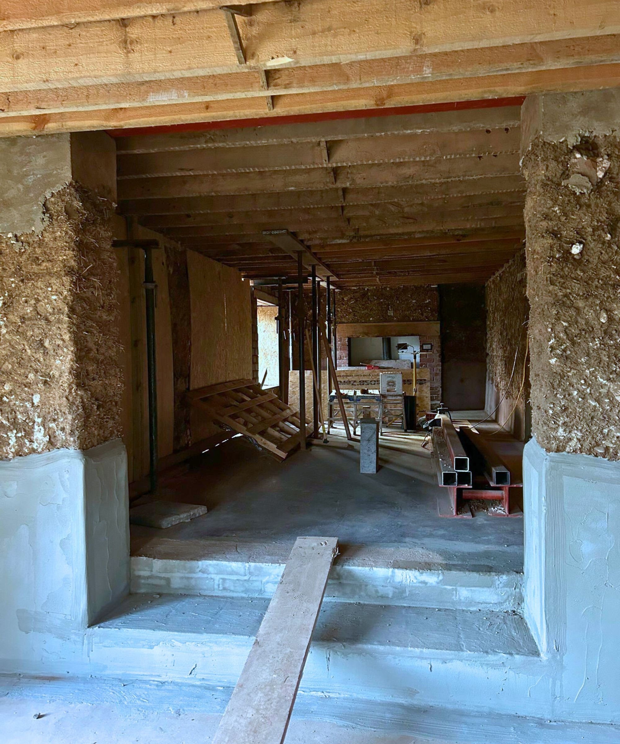 A large open space mid-renovation, showing exposed timber ceilings, cob walls and newly rendered lower sections. Steel beams, acrow props, plywood panels and scattered tools fill the room, while a temporary plank bridge spans the fresh concrete steps at the entrance.