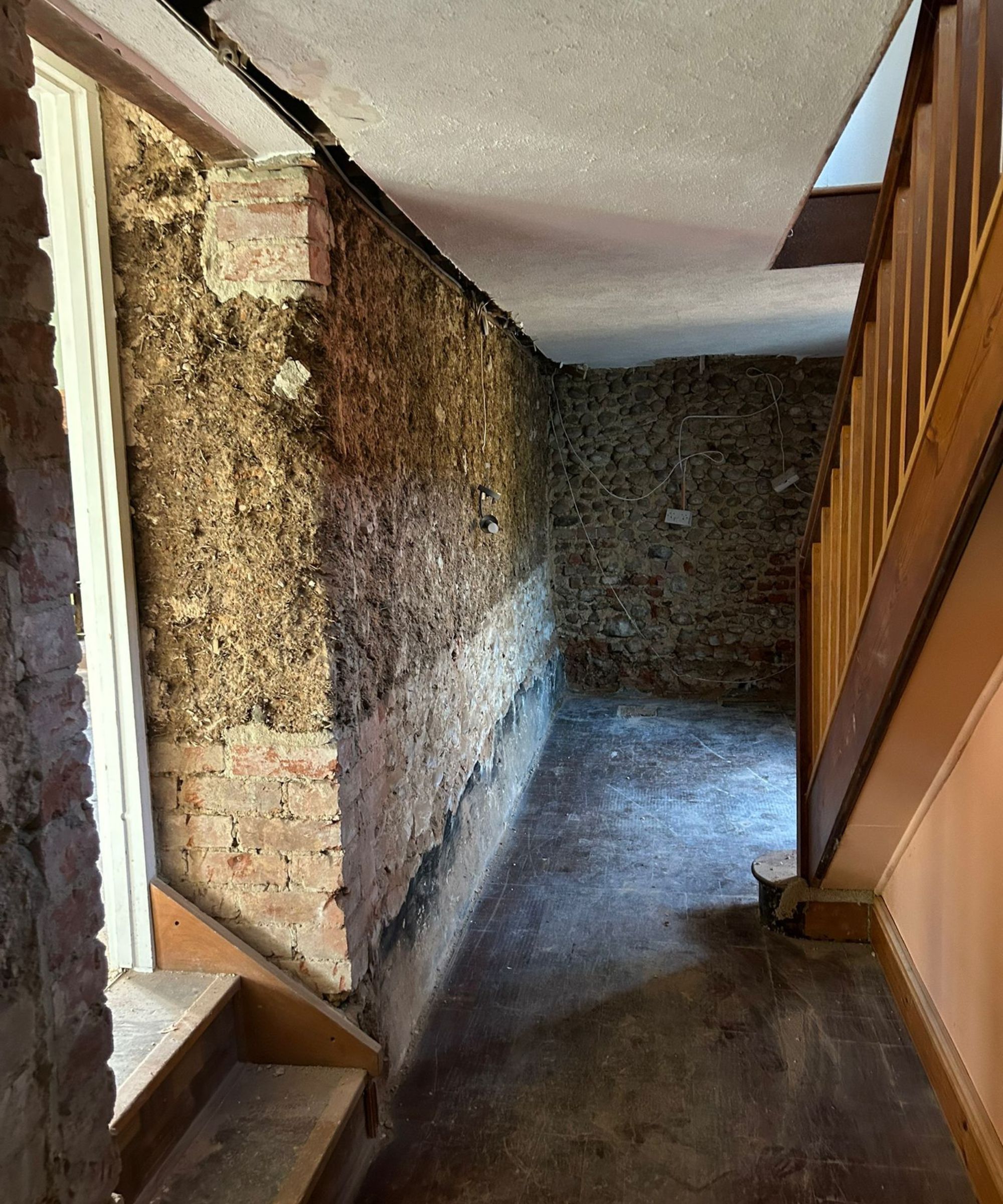 A stripped-back hallway with exposed brick and cob walls, visible wiring and a partially removed ceiling. The floor is bare, and the wooden staircase remains intact. The space has a raw, mid-renovation feel with structural layers revealed.