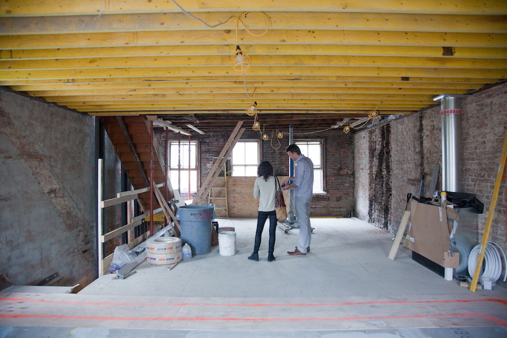 Two people review renovation plans in an unfinished brownstone interior with exposed beams and construction materials.