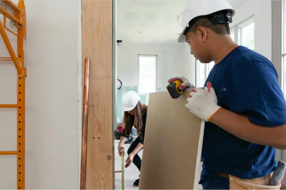 Two contractors wearing hard hats measure drywall inside an active renovation site.
