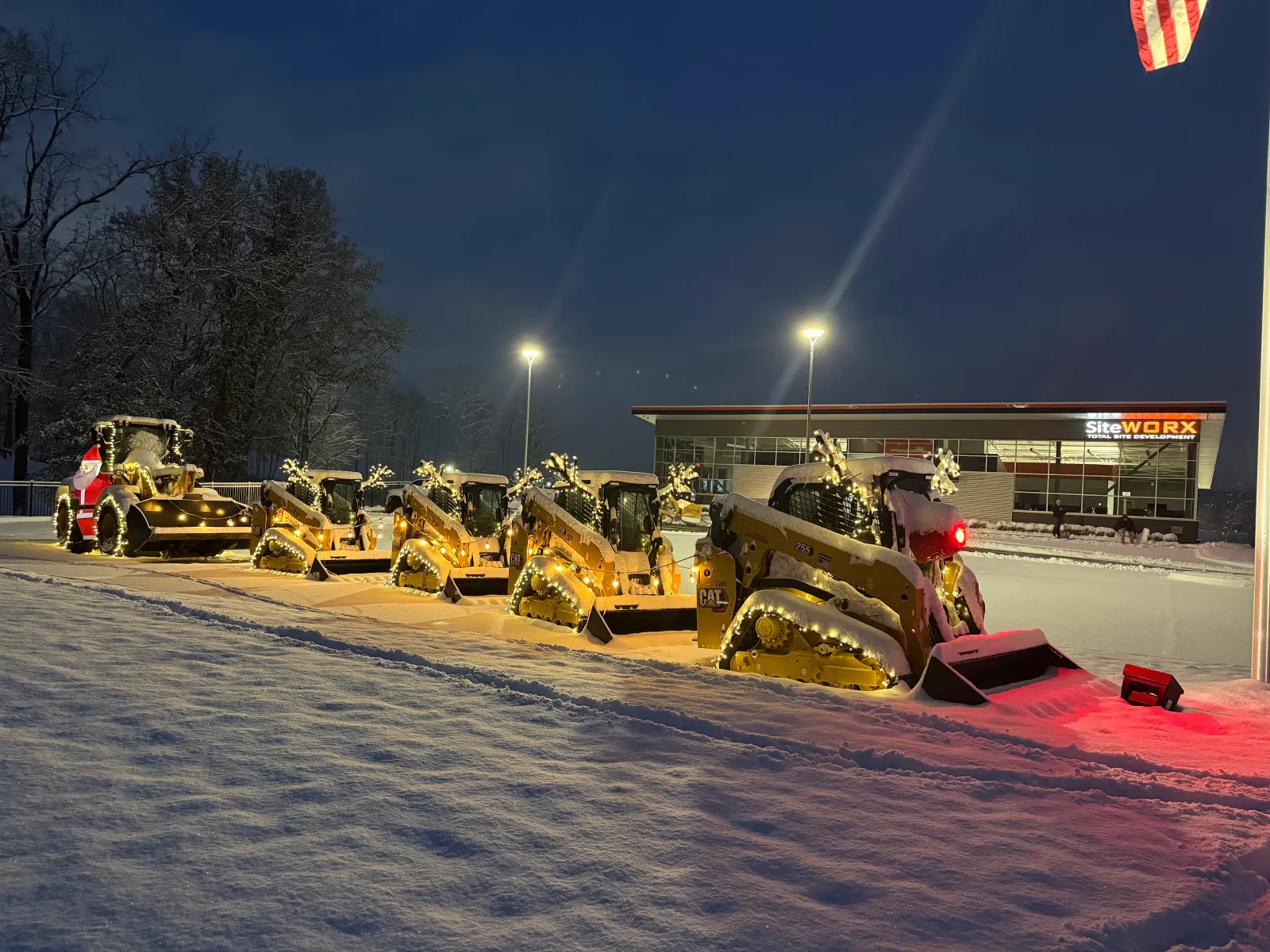 An inflatable Santa stands in front of a large wheel loader and several smaller pieces of heavy machinery, all adorned with reindeer antlers and lights. The lead piece of equipment has a glowing red "nose."