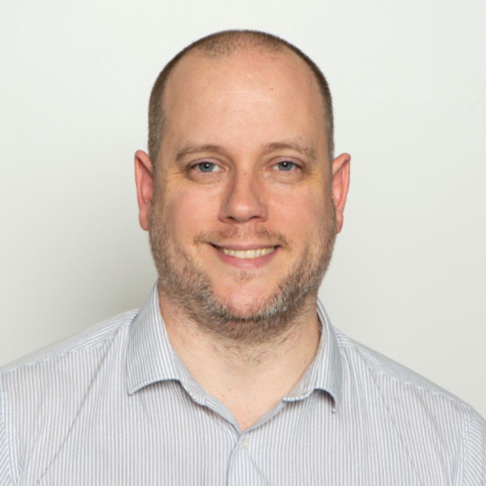 A man with a shaved head smiles warmly against a plain light background. He wears a light striped shirt and has a short beard. The portrait has a friendly, professional feel with soft, even lighting.