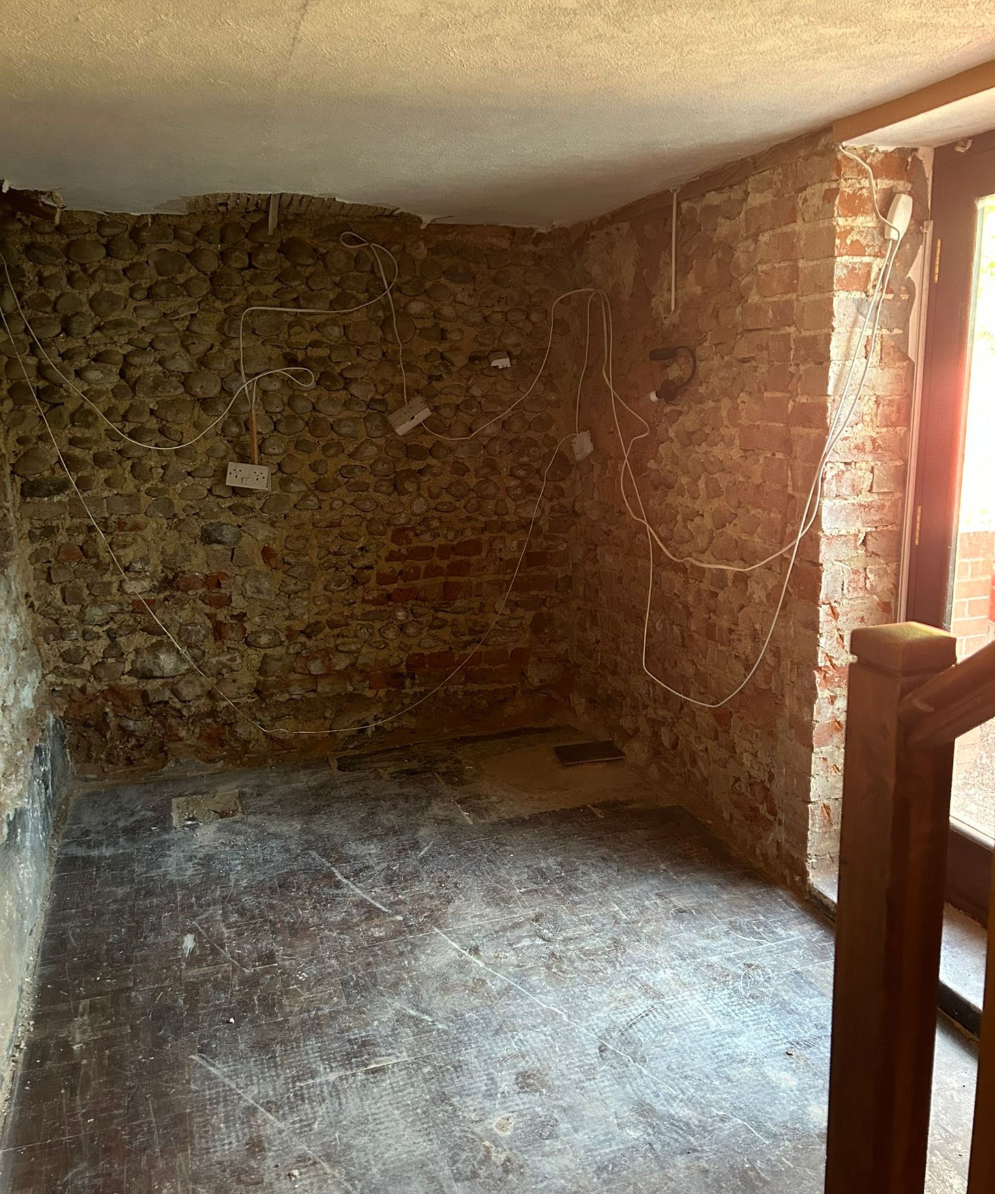 A bare room with exposed flint and brick walls, loose electrical cables running across the surface and a damaged ceiling edge. The floor is dusty and unfinished, while light enters through a glass door beside a wooden staircase.