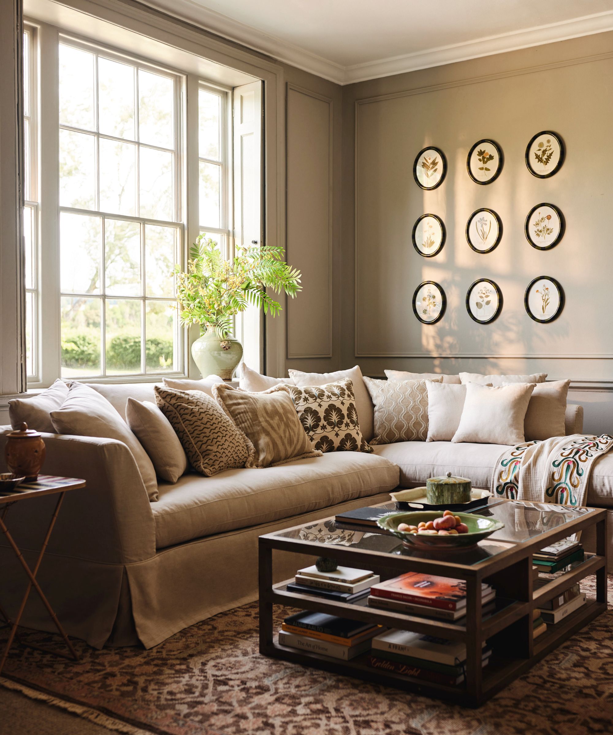 A serene, elegant living room with a large pale sectional sofa layered in patterned neutral cushions. Sunlight pours through tall sash windows, illuminating botanical artwork on the wall.