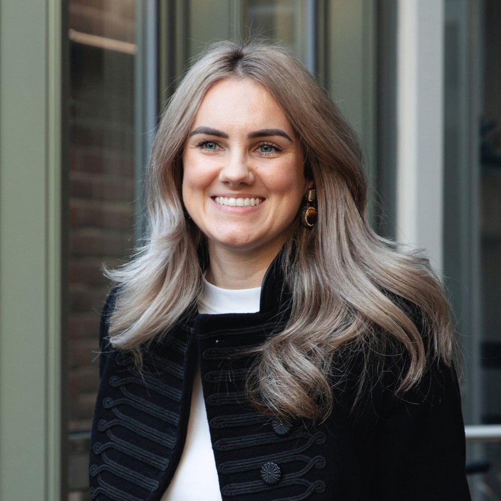 A woman with long, wavy blonde hair smiles while standing outdoors beside modern glass doors. She wears a black textured jacket and statement earrings. The portrait feels fresh, confident and natural, with soft daylight brightening her features.