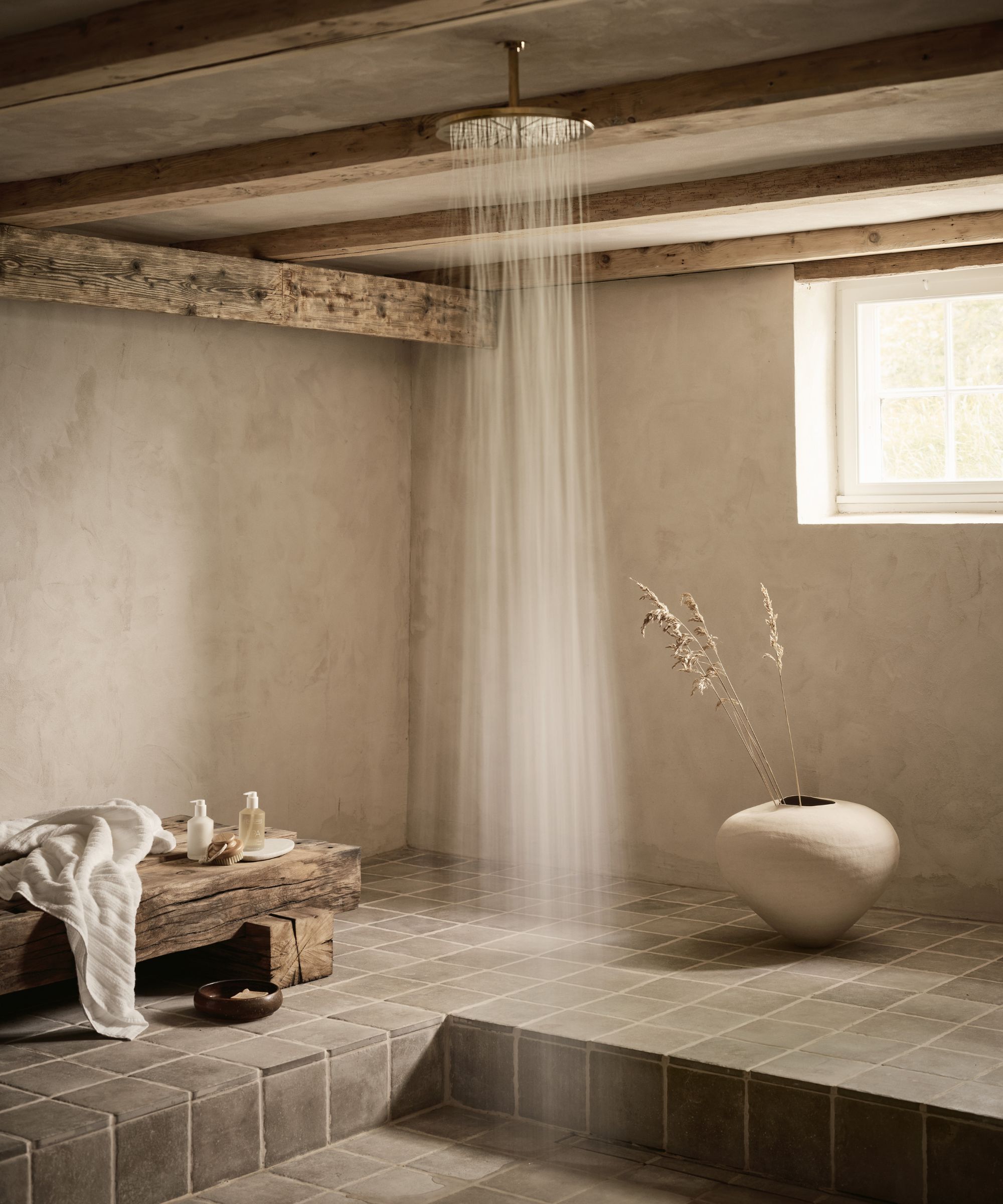 A rustic, spa-inspired wet room with tadelakt-style walls, exposed timber beams and a rainfall shower set over soft grey tiled flooring.