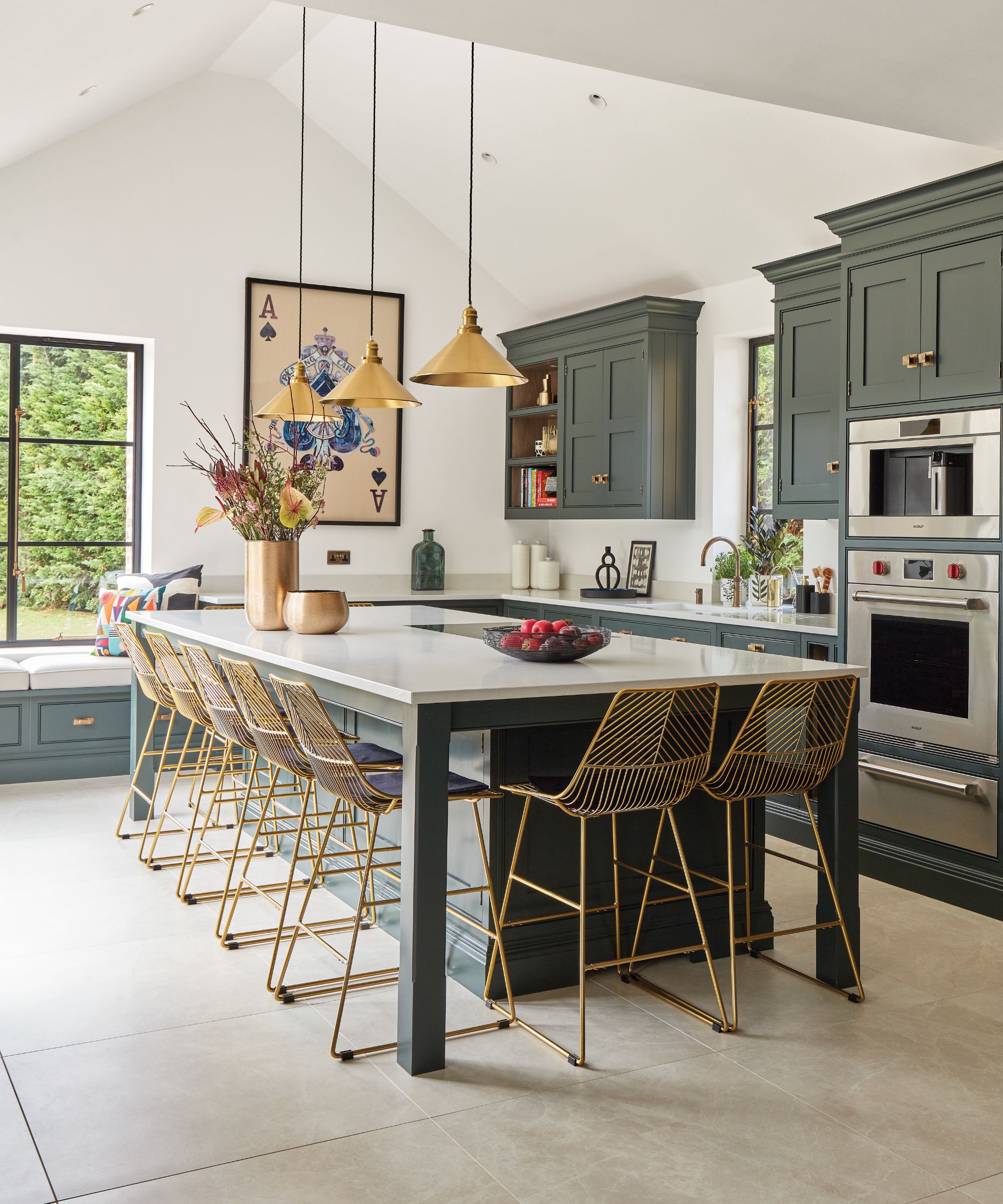 Large kitchen island with dark green cabinetry, white worktop and gold wire bar stools, lit by brass pendant lights in a bright, modern kitchen.