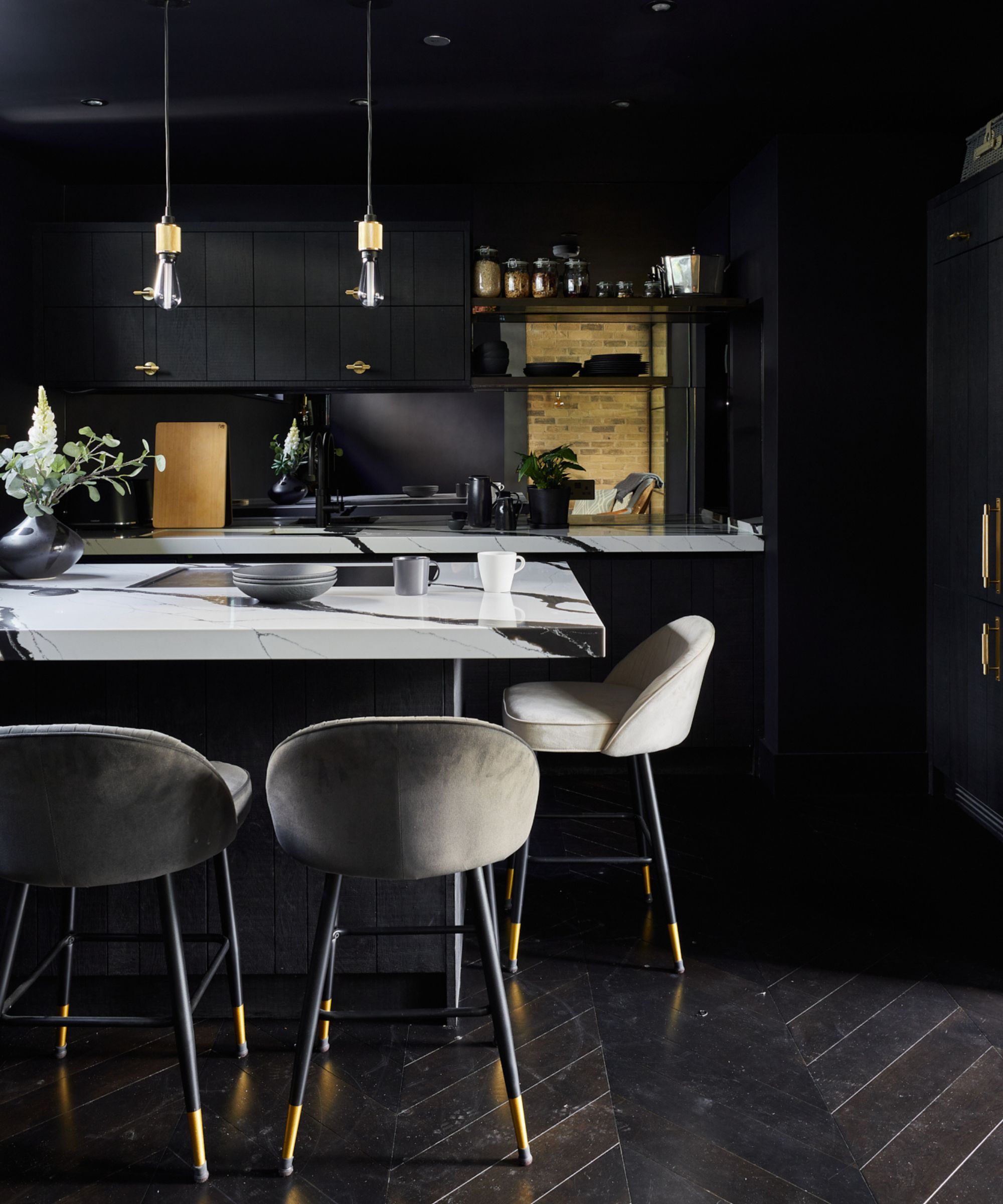 Dark, moody kitchen with black cabinets and marble island. Three upholstered bar stools sit beneath slim pendant lights, with open shelving and brick detail behind.