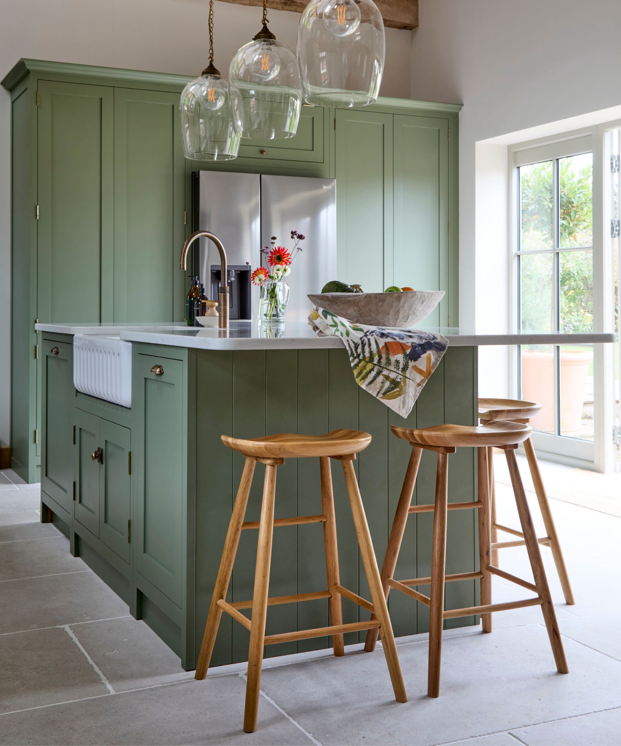 Light green kitchen island with panelled sides and white worktop, paired with three wooden bar stools beneath glass pendant lights by a window.
