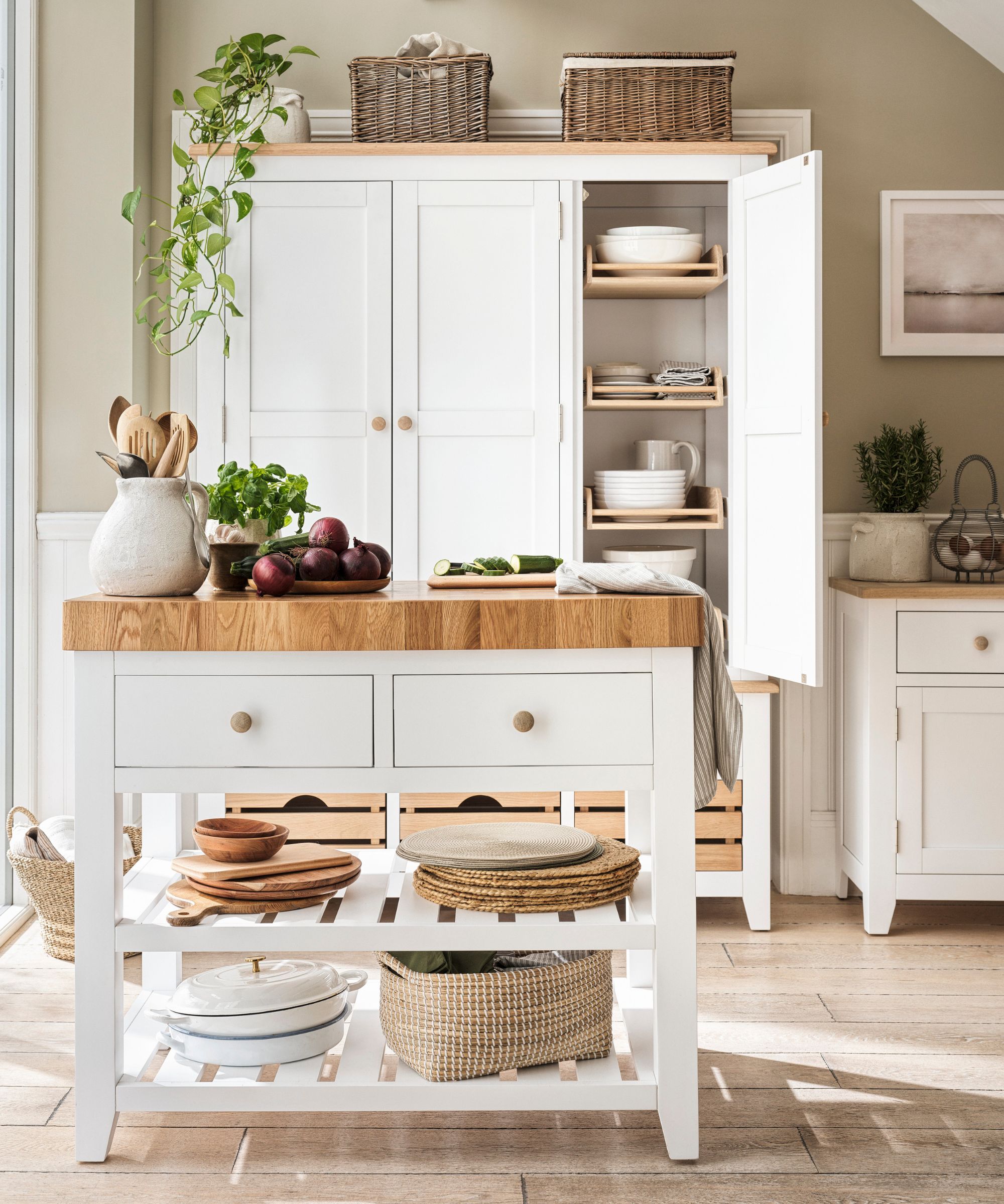 White portable kitchen island with thick wooden worktop, open shelves and baskets, styled with vegetables and crockery in a light, country-style kitchen.