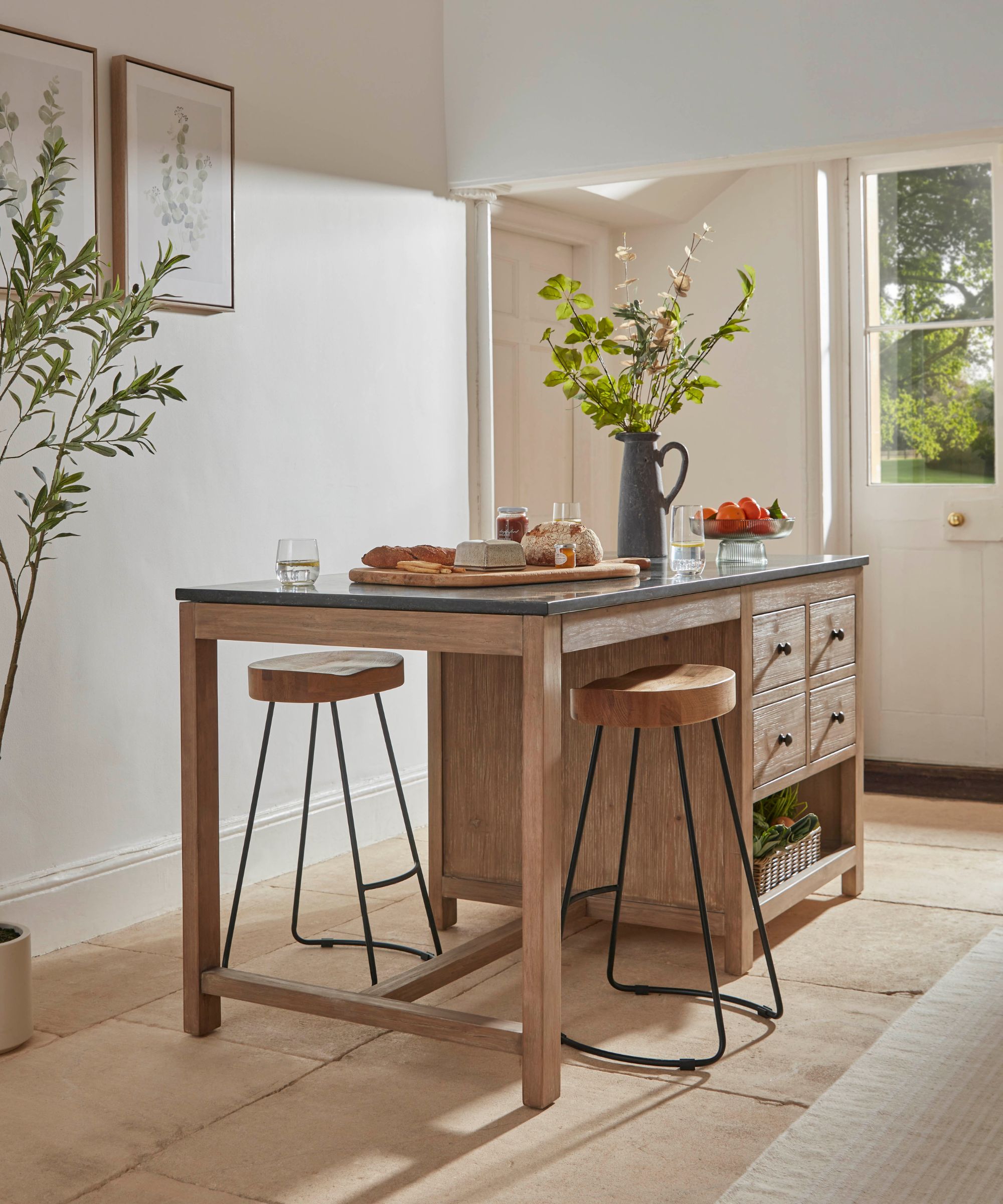 Wooden portable kitchen island with black worktop and stools, styled with bread, jam, flowers and glasses in a bright, traditional kitchen.