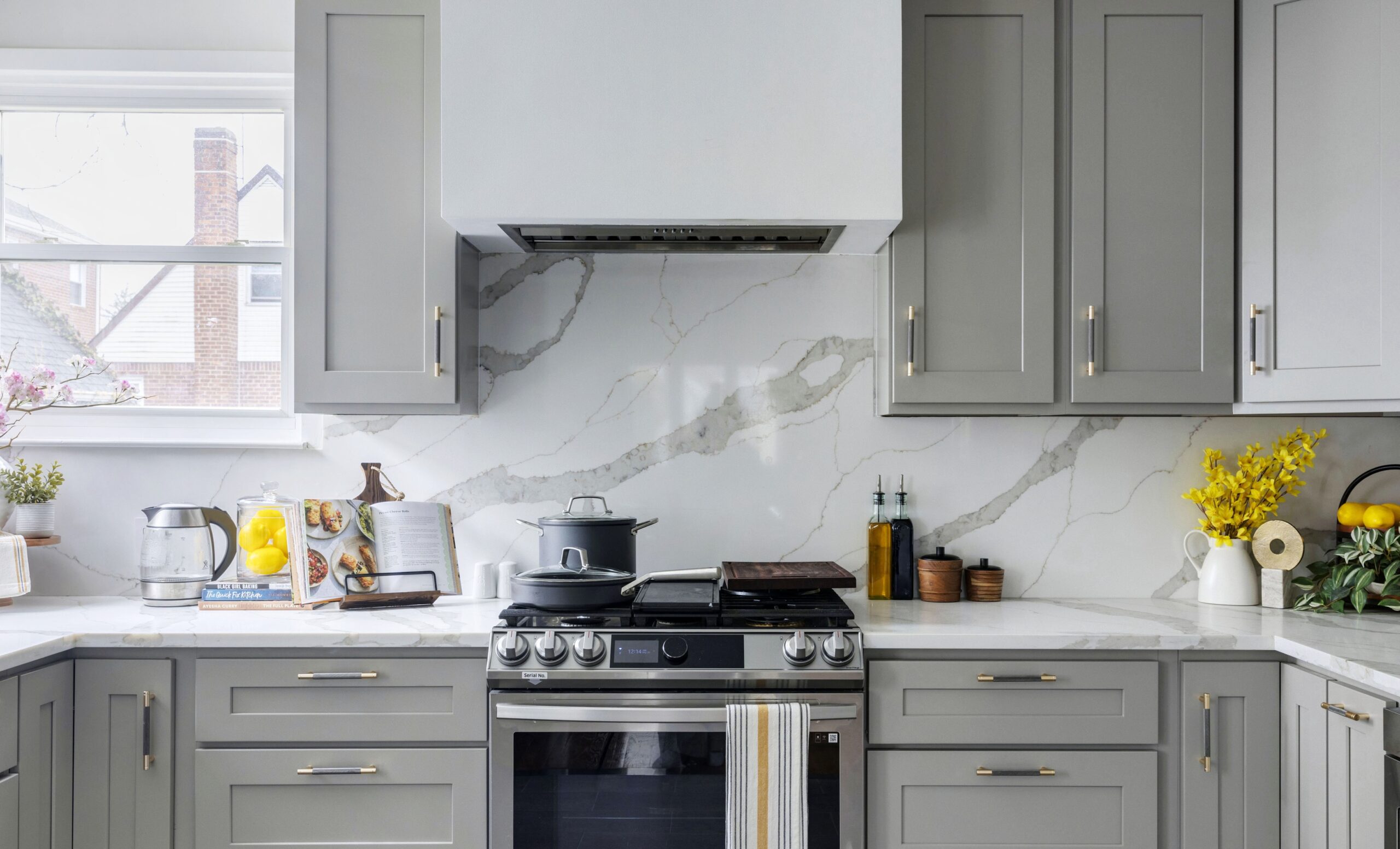Backsplash slab shows sweeping grey veins, set behind a stainless range, grey shaker cabinetry, brass pulls, and a crisp white hood at center near window.